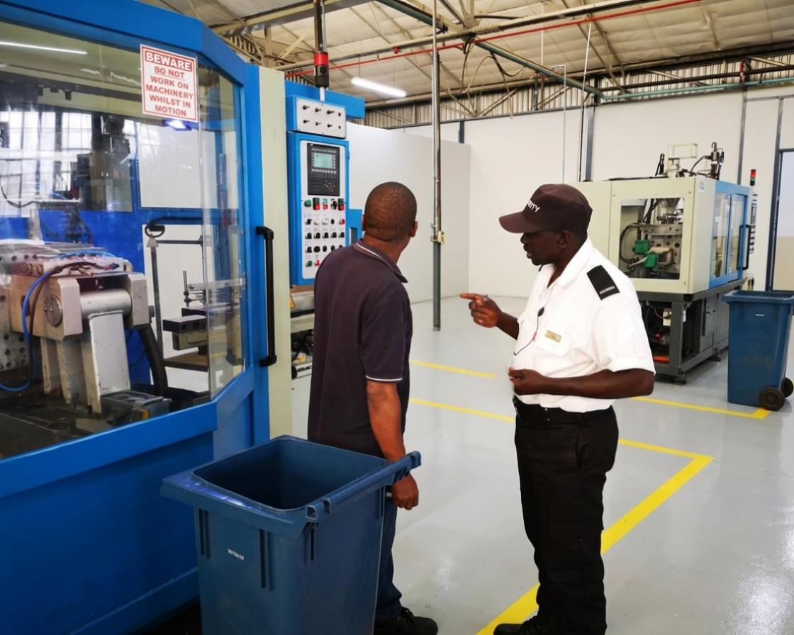 A uniformed security guard wearing a black 'SECURITY' cap and a white short-sleeved shirt is conducting an inspection or safety brief