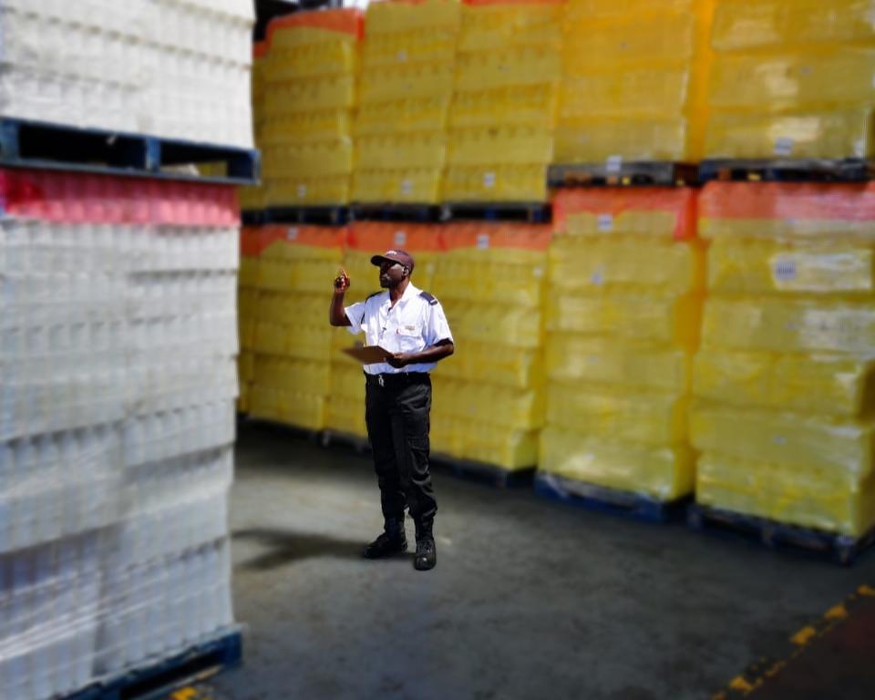 A professional security guard in a white uniform and cap performing an inventory inspection in a large warehouse with stacked pallets of goods.