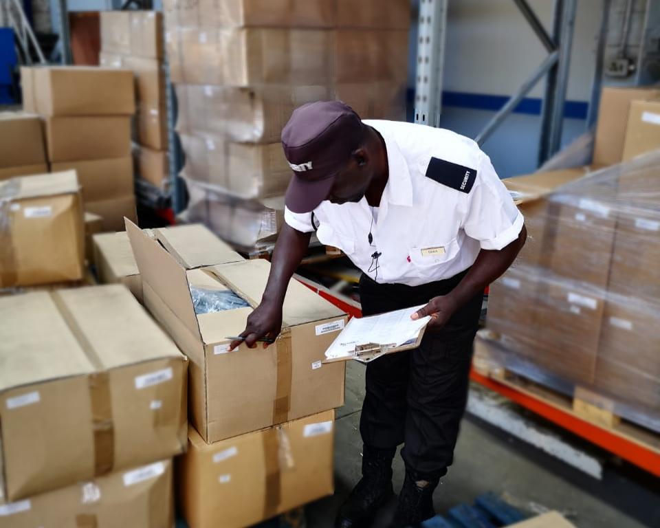 A security guard in a professional uniform conducting a detailed stock inspection and inventory check on palletized boxes in a warehouse.